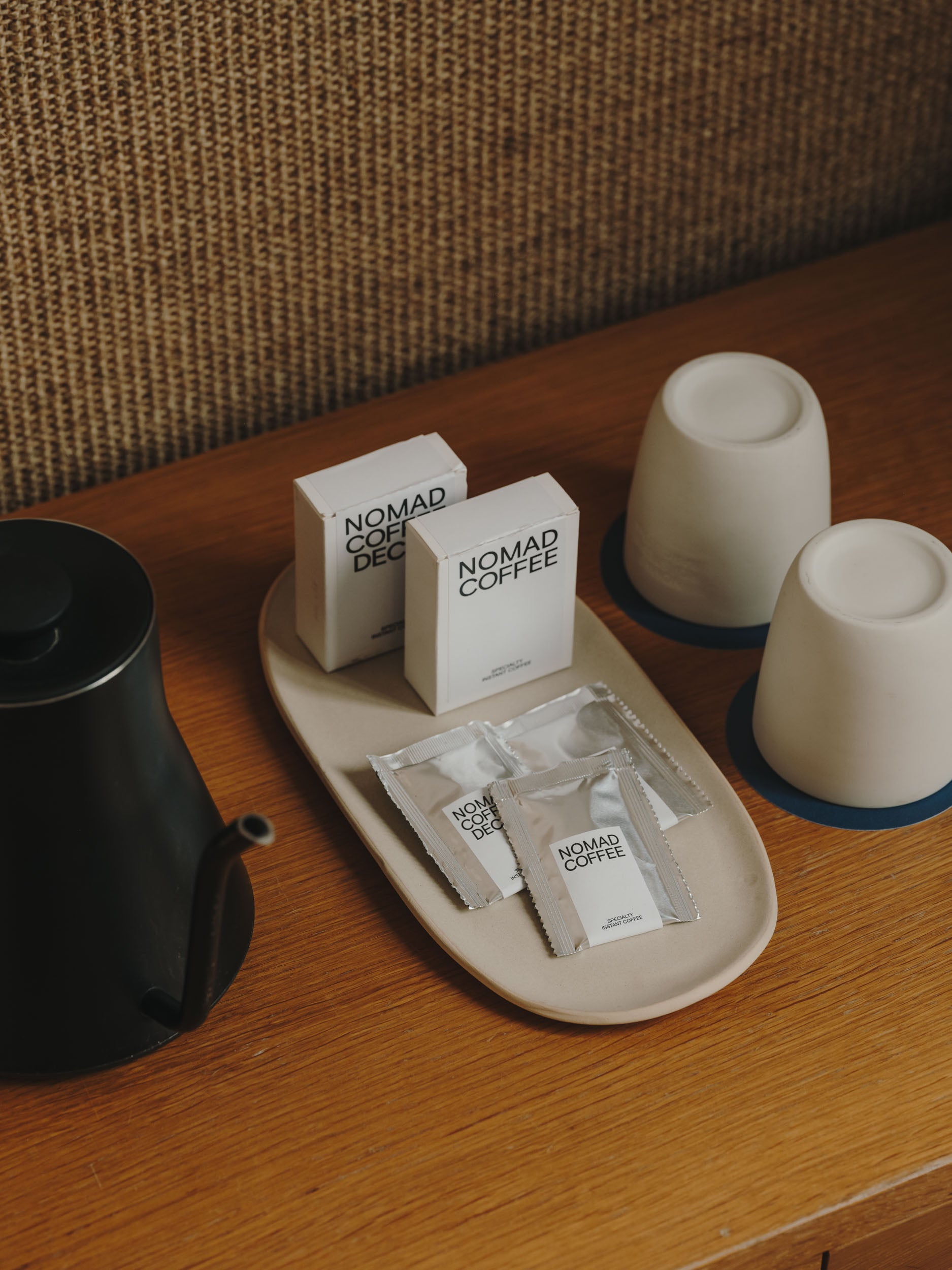 Set de cortesía sobre una mesa de madera que incluye una tetera negra, dos tazas blancas invertidas, y un plato con sobres monodosis de café NOMAD COFFEE —tanto normal como descafeinado— y dos cajas con el mismo branding.
