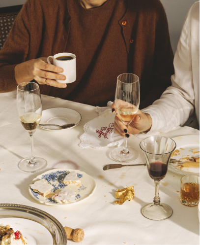 Mesa tras una comida, con platos con restos de postre, copas de vino y cava, una taza de café y manos de dos personas en primer plano. Una lleva un jersey marrón y sostiene la taza; la otra viste de blanco y levanta una copa. La mesa está cubierta con un mantel blanco.