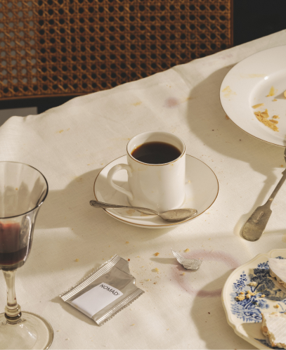 Taza de café sobre un platillo con una cuchara, en una mesa con mantel manchado y restos de comida. Hay una copa con vino tinto, platos con migas y un sobre individual de café NOMAD abierto.