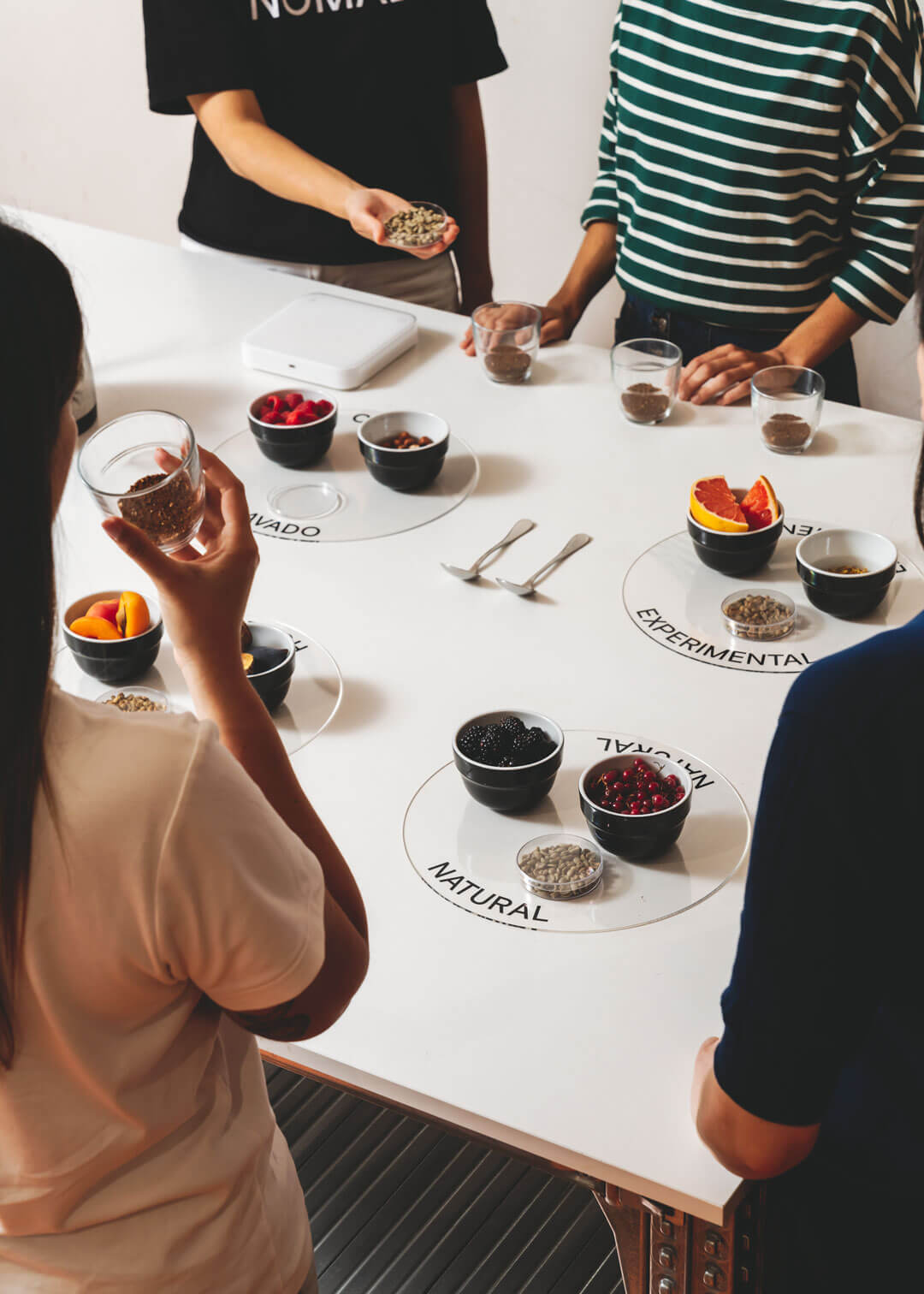 Cinco personas alrededor de una mesa blanca con muestras sensoriales. Cada grupo incluye una taza con café molido y pequeños boles con frutas o ingredientes representativos: frutos rojos, naranja, albaricoque, etc. Las categorías “LAVADO”, “NATURAL” y “EXPERIMENTAL” están marcadas en la mesa, indicando tipos de procesos de café.