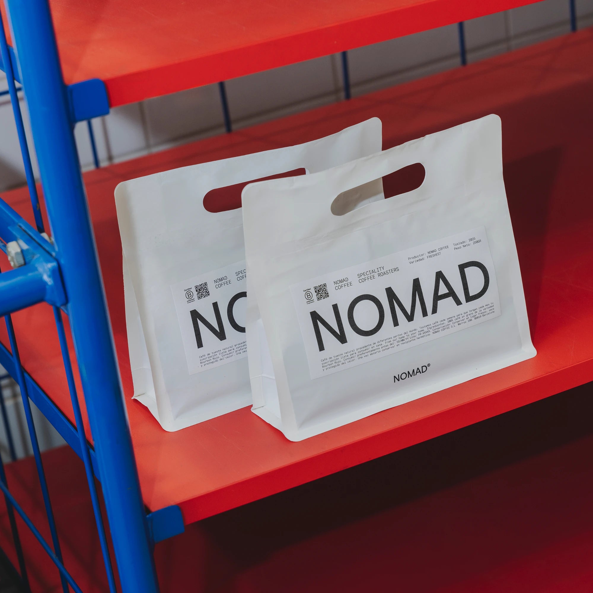 Dos bolsas blancas de café de Nomad con diseño impreso y asas troqueladas, colocadas sobre una estantería roja con estructura metálica azul.