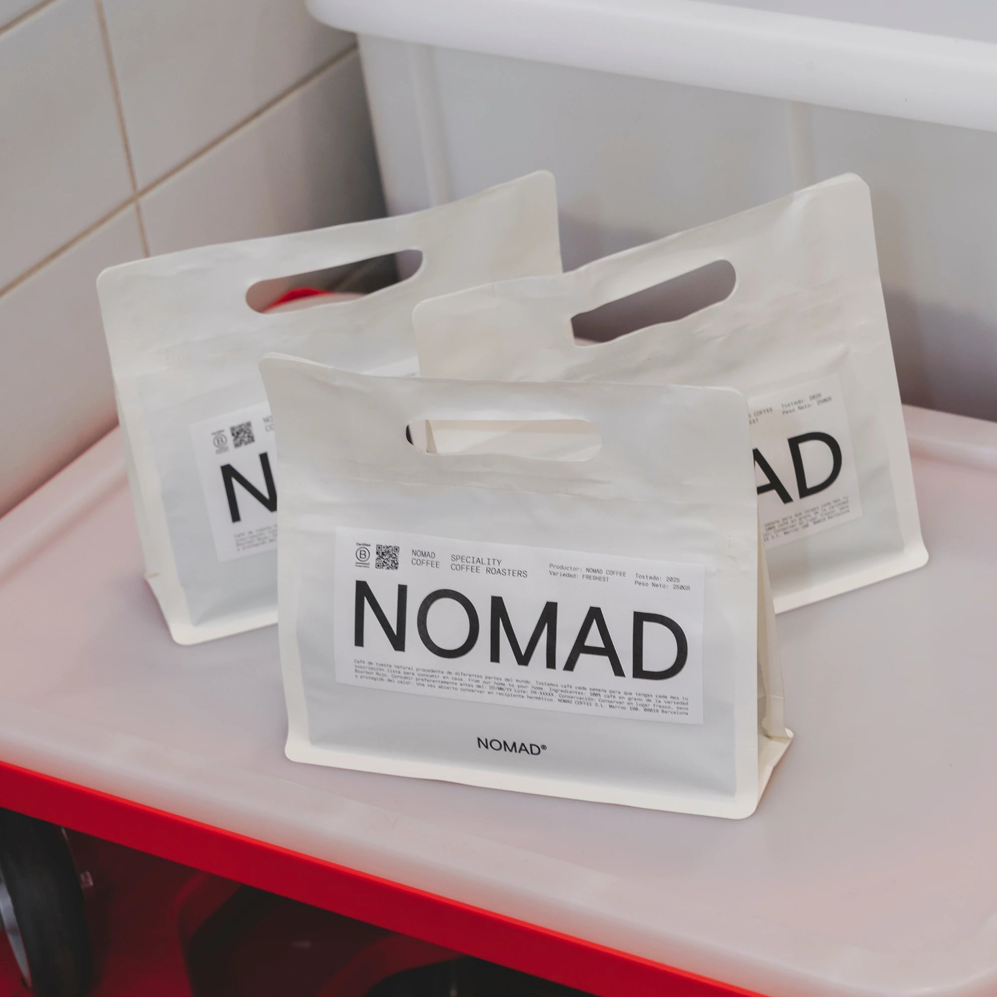 Tres bolsas blancas de café de Nomad con diseño impreso y asa troquelada, colocadas sobre una bandeja roja en un entorno de trabajo.