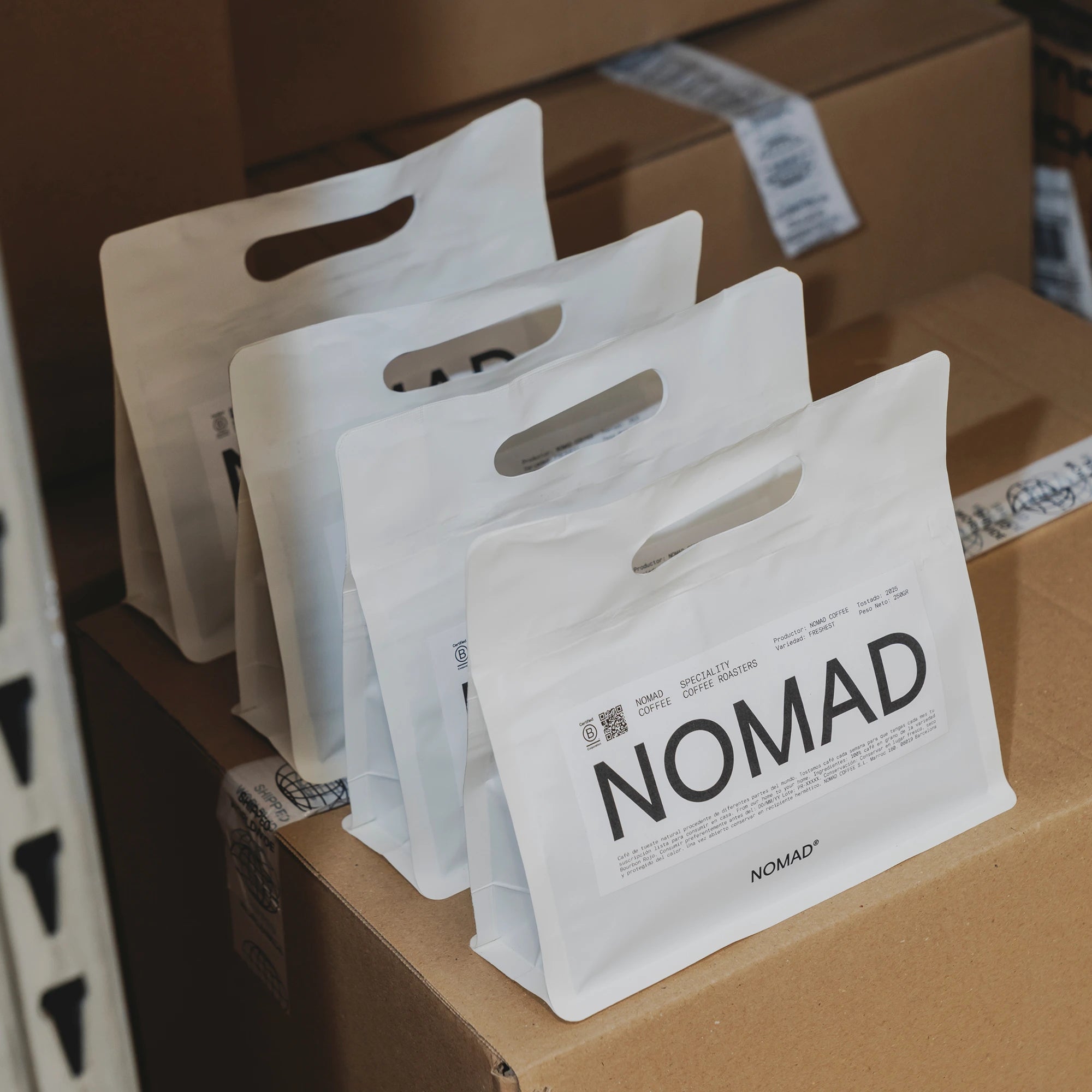 Four white Nomad coffee bags with die-cut handle and printed design, placed on cardboard boxes ready for shipping.