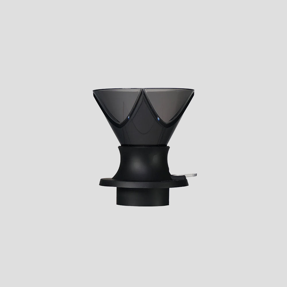 Front view of a modern design coffee dripper in black, with an inverted conical shape and geometric details. It is made of dark translucent plastic on top and a more opaque sturdy base with a side lever, on a light gray background.