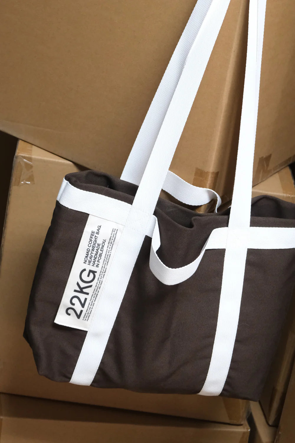 Bolsa tote de Nomad Coffee de color marrón oscuro con asas blancas y etiqueta visible “22KG”, apoyada sobre cajas de cartón marrón.